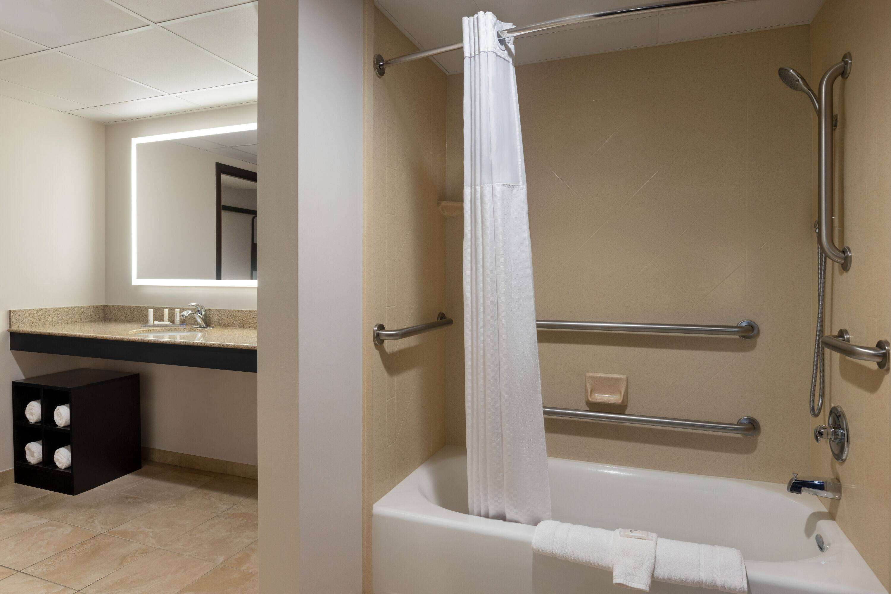 A modern bathroom with a bathtub, a handrail, a towel rack, and a mirror.