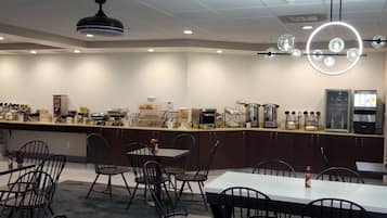 A modern breakfast bar with a variety of food and beverage options, including a coffee machine and a refrigerator. There are several tables and chairs arranged for dining.