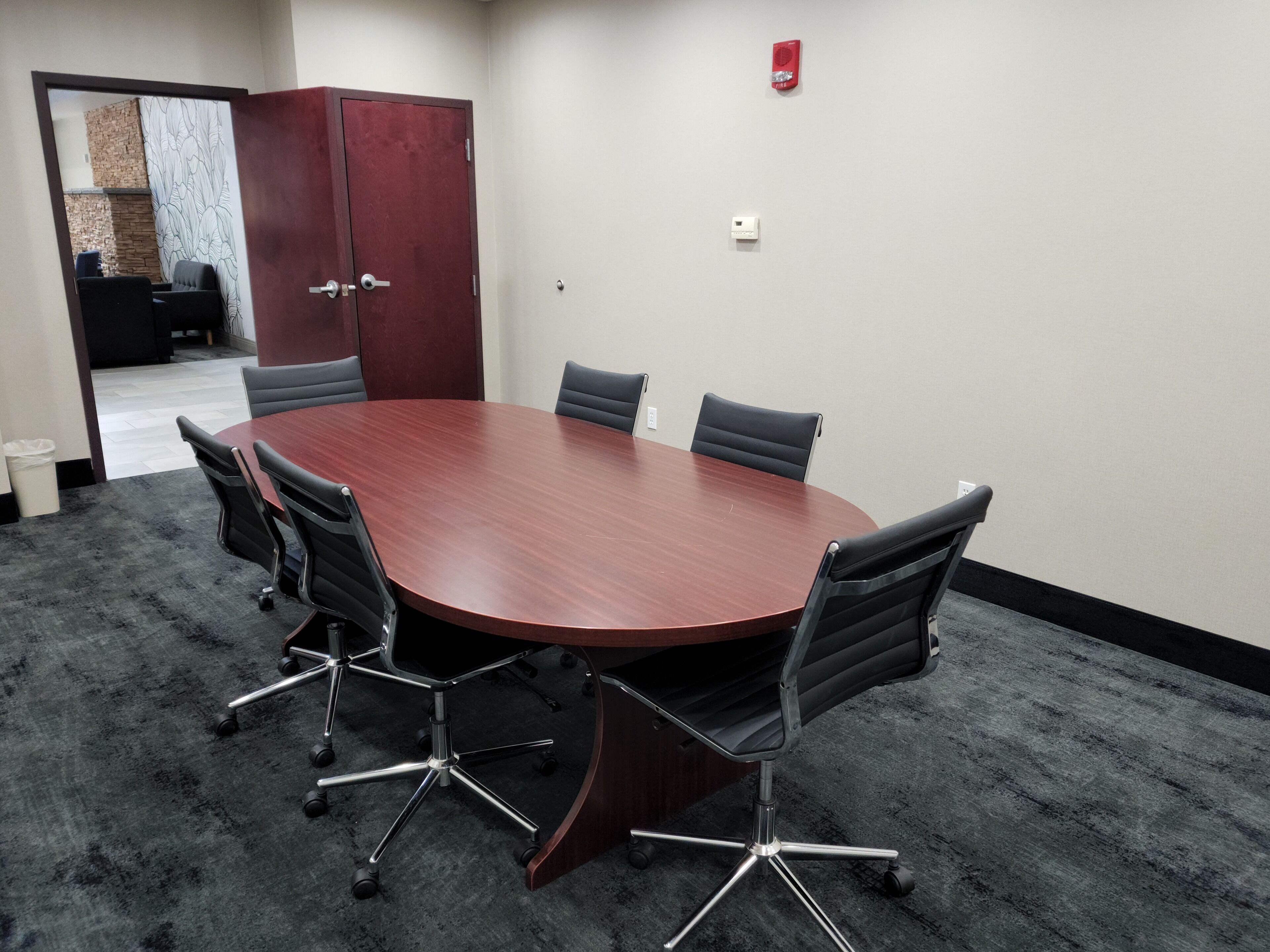 A conference room with a round table, six chairs, a red cabinet, and a fire alarm on the wall.