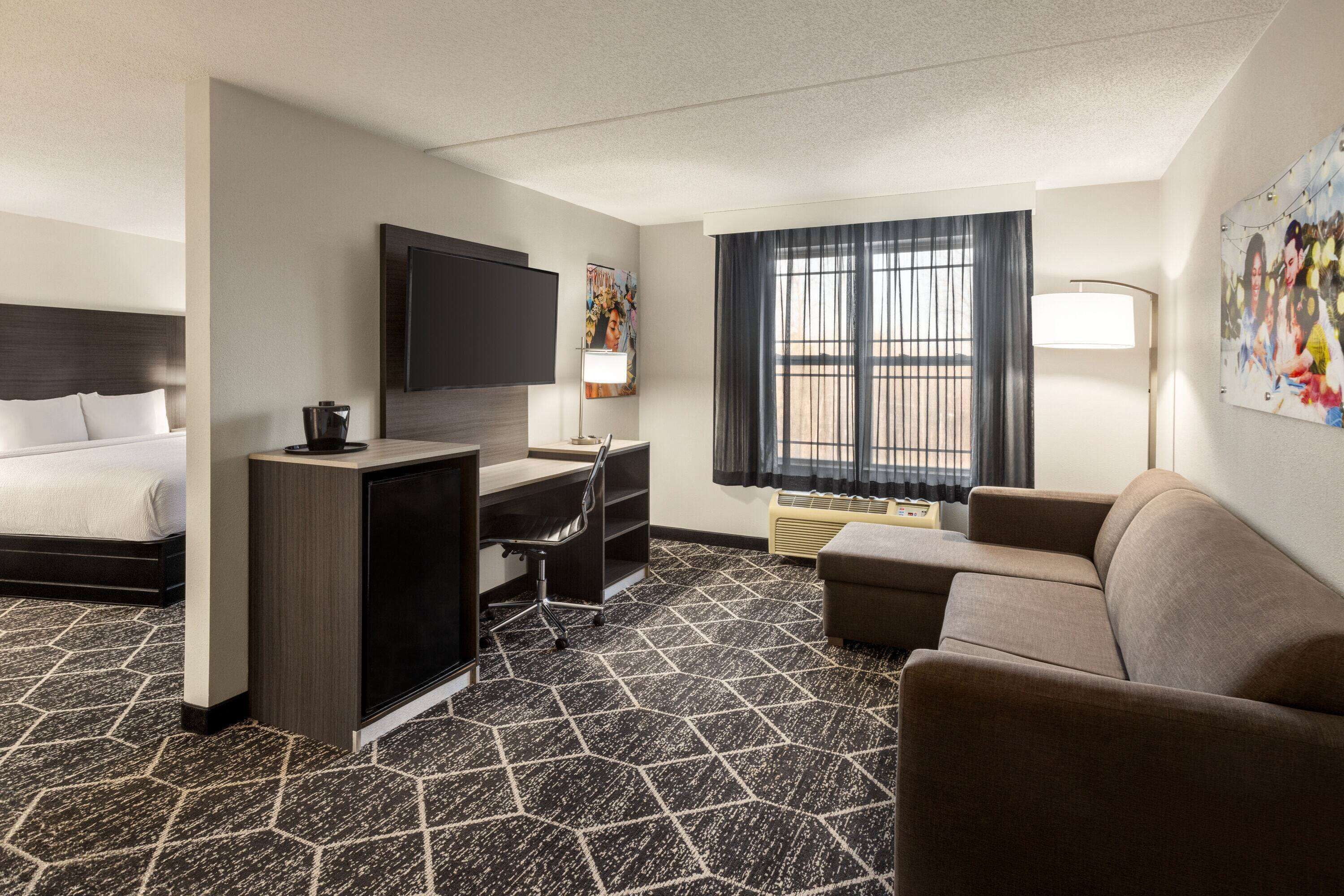 Habitación de hotel con cama, escritorio con silla, sofá, televisor y ventana con cortinas.