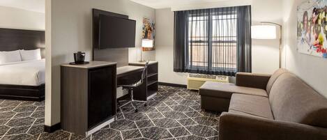 Habitación de hotel con cama, escritorio con silla, sofá, televisor y ventana con cortinas.