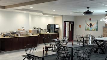 Un comedor con mesas y sillas, una zona de cocina con electrodomésticos, y un cuadro en la pared.