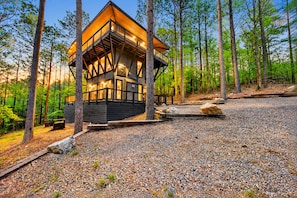 A cabin with a wooden exterior, a balcony, and a gravel ground. - 3 Story Treehouse | Views, Pool Table, Hot Tub (Broken Bow)