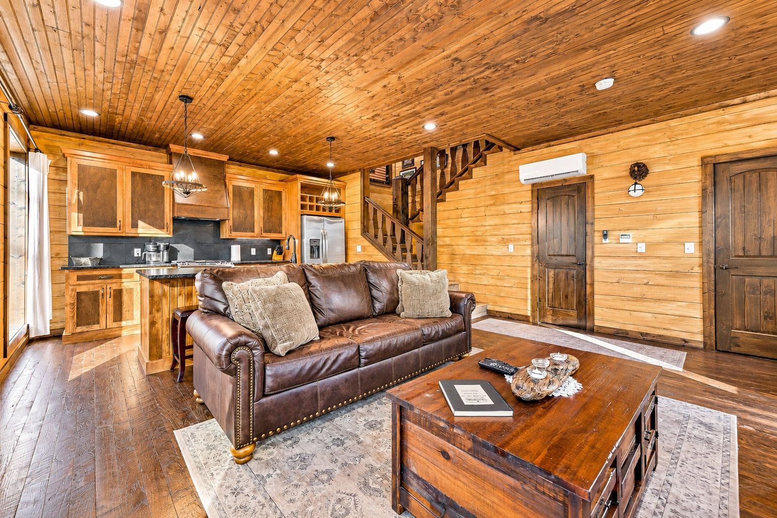 A cozy cabin interior with a leather sofa, wooden flooring, a kitchen area, and a staircase.