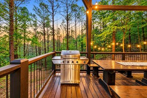 An outdoor deck with a stainless steel grill, wooden benches, and string lights. - 3 Story Treehouse | Views, Pool Table, Hot Tub (Broken Bow)