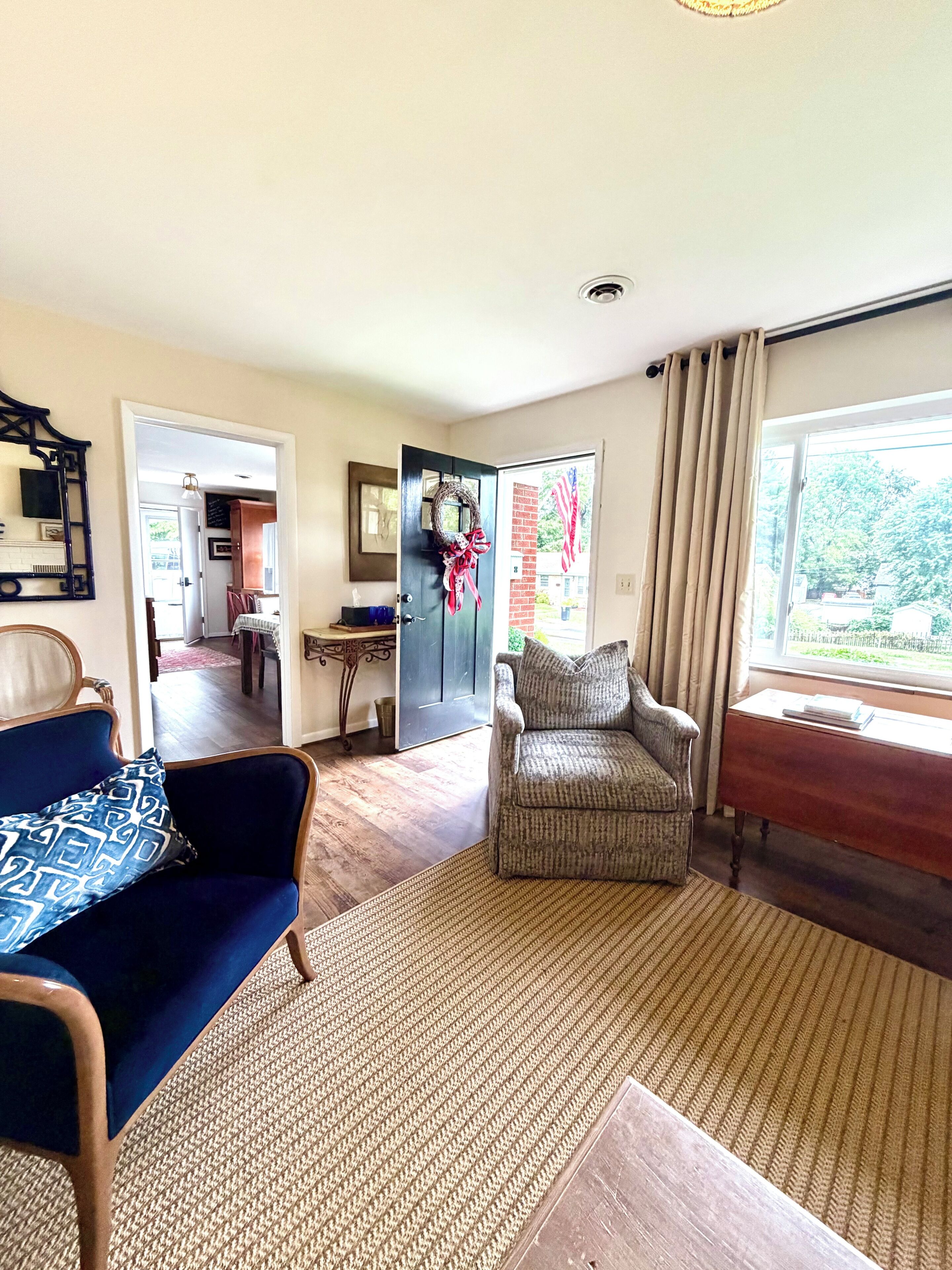 A living room with a blue sofa, a patterned armchair, and a wooden coffee table. There is a door leading to another room, and a window with curtains.