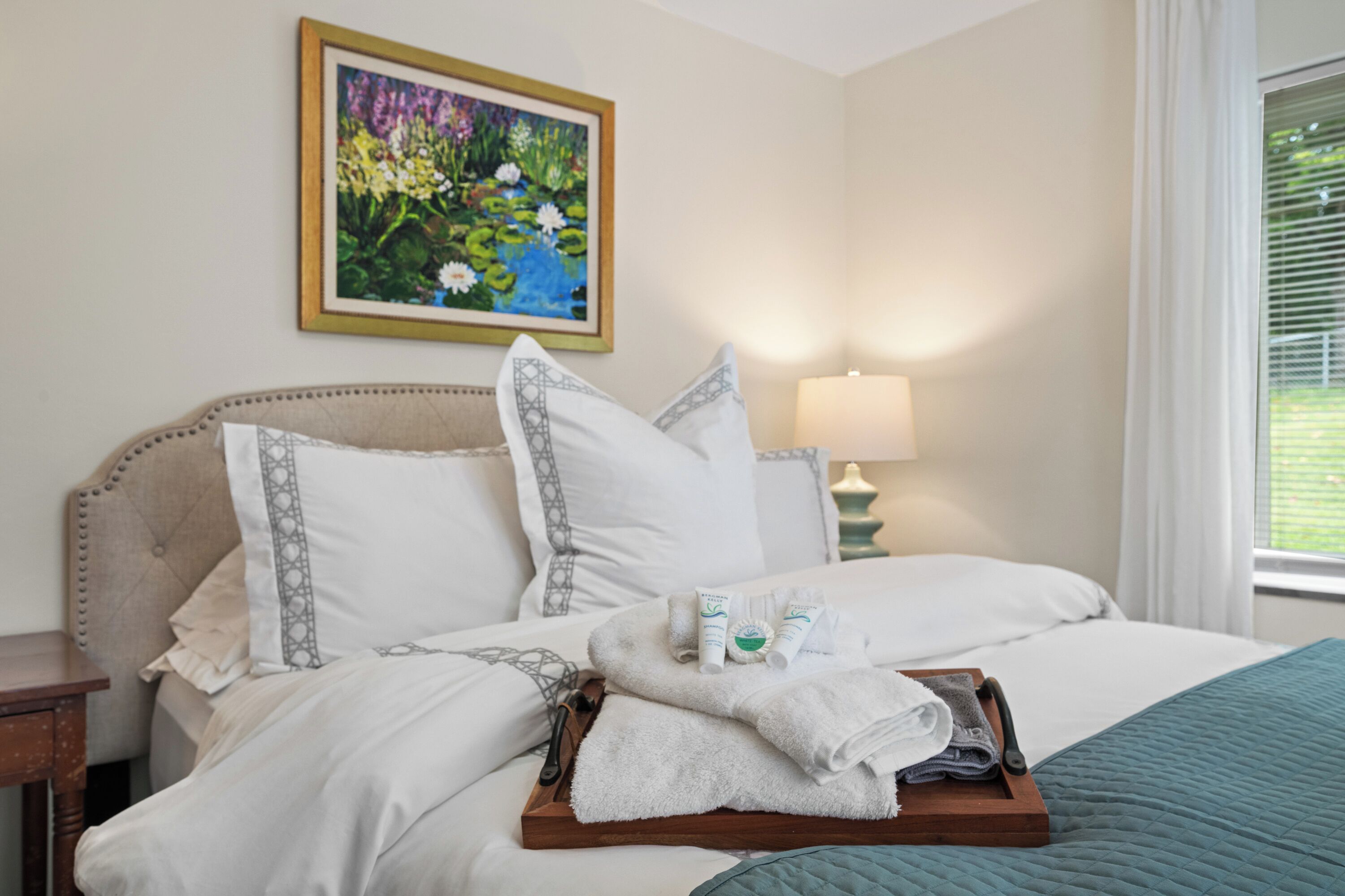 Una cama bien hecha con sábanas blancas, una bandeja de madera con toallas y artículos de aseo, y un cuadro de flores en la pared.