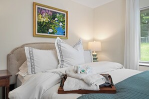A neatly made bed with white linens, a wooden tray with towels and toiletries, and a painting of flowers on the wall.