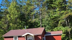 Une petite cabane en bois rouge, avec un toit en métal, une porte d’entrée et un petit porche.