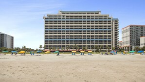 Een hotel met meerdere verdiepingen en uitzicht op het strand, met diverse balkons en een zandstrand voorzien van parasols en ligstoelen.