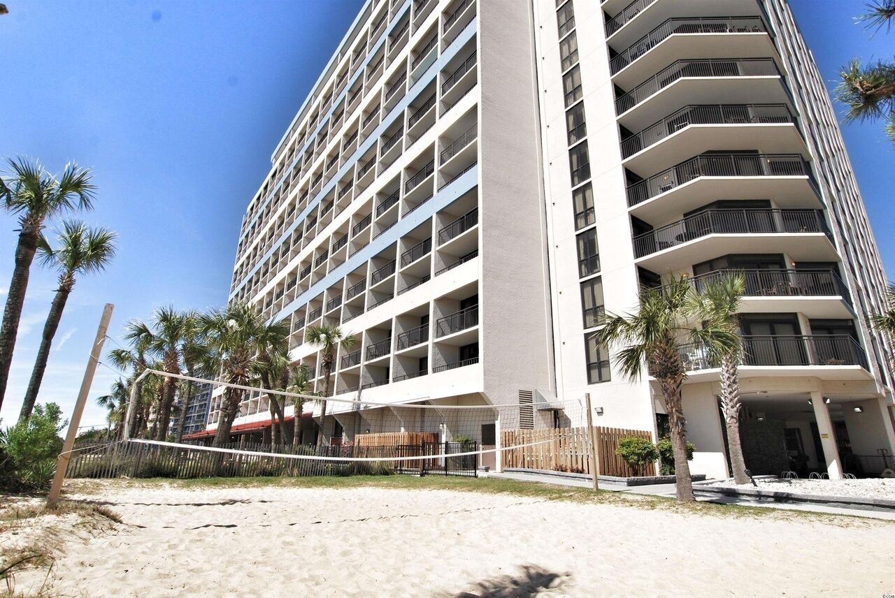A high-rise hotel with multiple balconies, a sandy beach in front, and palm trees.
