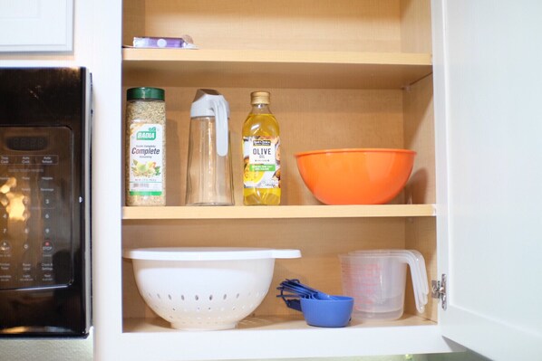 A kitchen cabinet with a microwave, a colander, a measuring cup, a bottle of olive oil, a container of seasoning, and an orange mixing bowl. - Peaceful 3-2 Home w/ Pool, 4min to beach, RV P-king, BBQ, Firepit, Fast Internet (Gulf Breeze)