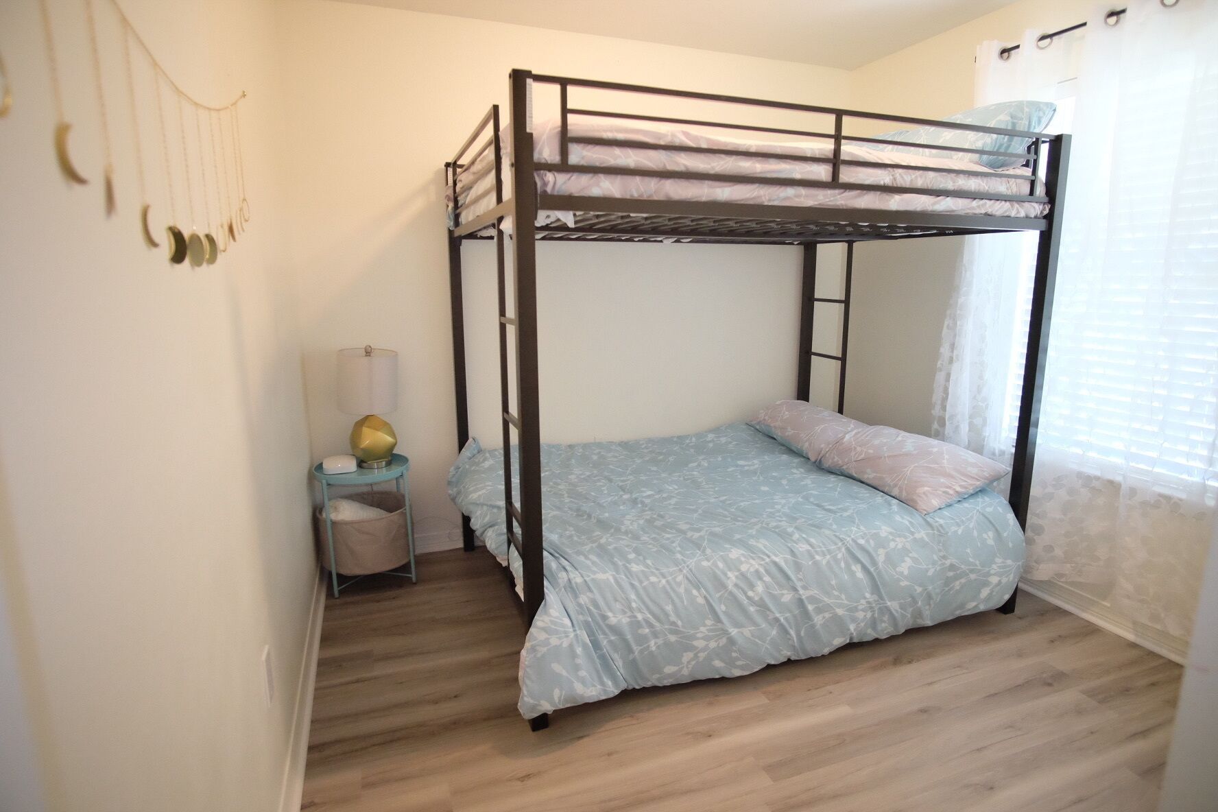 Quarto com beliches, piso de madeira, mesa de cabeceira com abajur e janela com cortinas.