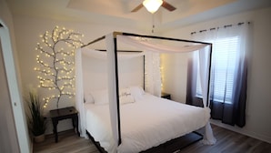A four-poster canopy bed with white bedding, a bedside table, a wall-mounted tree light, and a ceiling fan. - Peaceful 3-2 Home w/ Pool, 4min to beach, RV P-king, BBQ, Firepit, Fast Internet (Gulf Breeze)