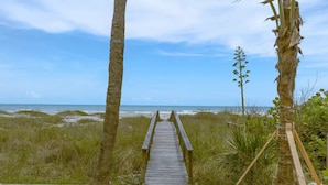 Une promenade en bois mène à une plage bordée de dunes et, sur la droite, on aperçoit un palmier.
