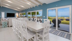 Un vaste espace de vie comprenant une grande table et des chaises blanches, un canapé et un téléviseur. De grandes fenêtres offrent une vue sur la plage.