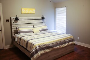 Une chambre à coucher comprenant un lit, une tête de lit, une fenêtre avec des stores et une œuvre d’art accrochée au mur.