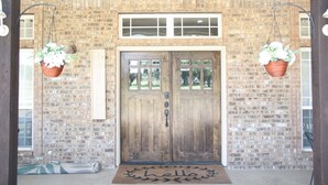 A wooden double door with a welcome mat, hanging flower baskets, and a brick wall.