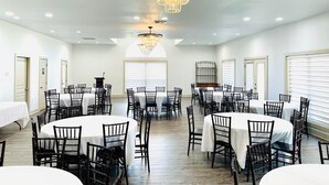A spacious dining area with round tables and chairs, a large chandelier, and large windows with blinds.