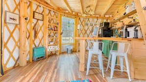 Une cabane en bois chaleureuse, dotée d’un coin cuisine, d’un espace repas avec des chaises blanches et d’un tapis coloré sur le sol.