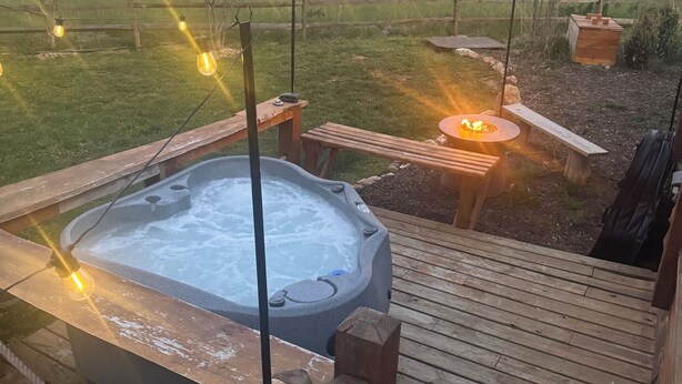 Un bain à remous sur une terrasse en bois, avec vue sur un champ verdoyant et une colline en arrière-plan.