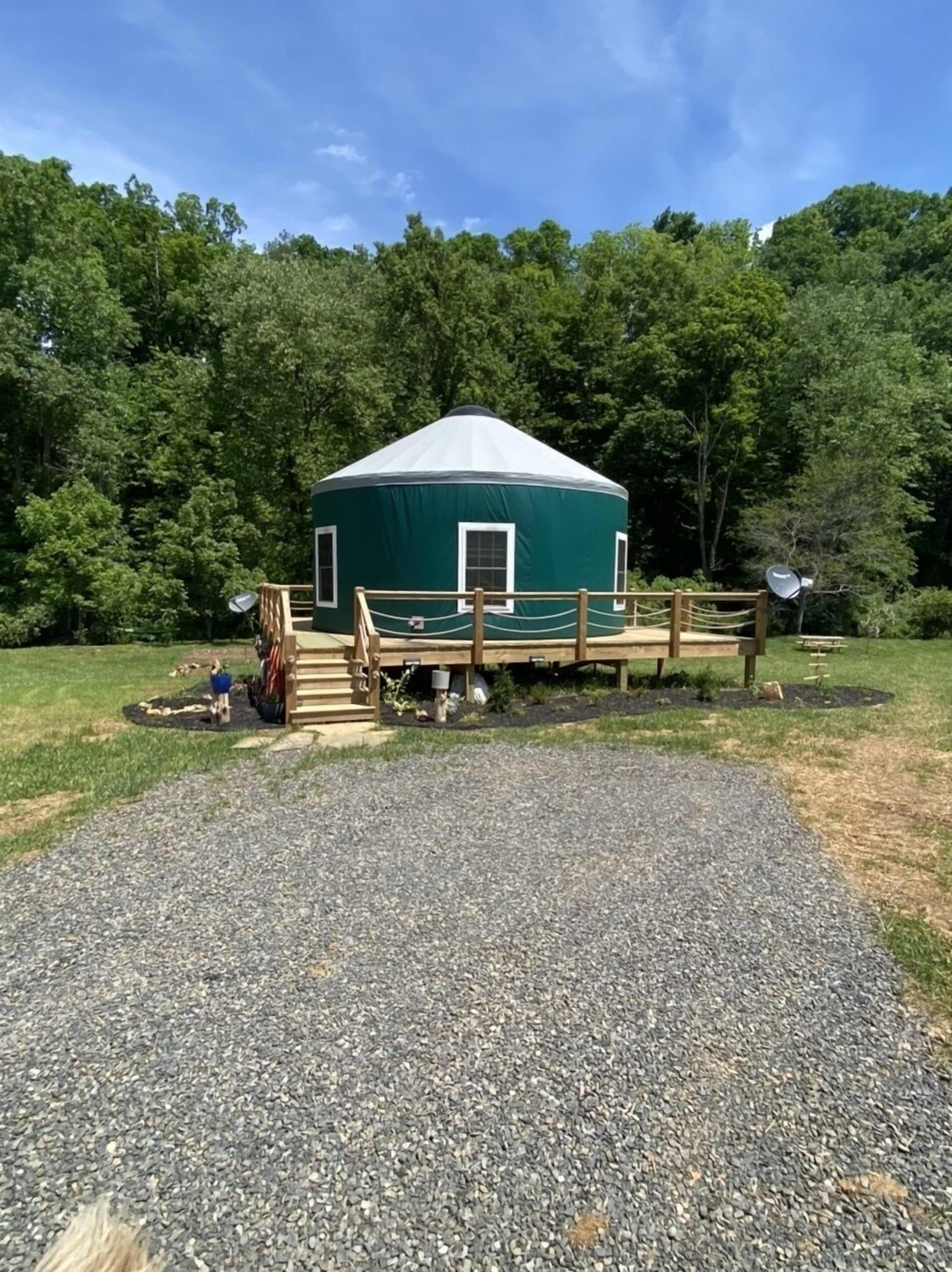 A green yurt with a white roof, surrounded by a wooden deck and gravel path, set in a forested area.