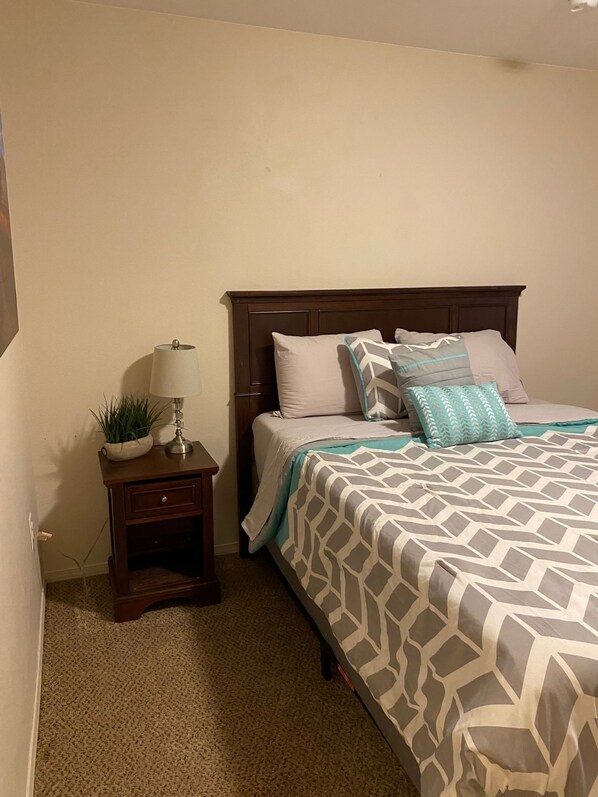 Un dormitorio con cama, mesita de noche, lámpara y una planta pequeña.