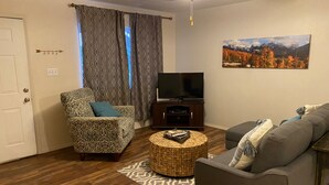 Ein Wohnzimmer mit einem gemusterten Sessel, einem grauen Sofa, einem Fernseher auf einem Holzboden und einem Gemälde von einer Berglandschaft.