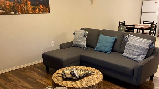 Ein modernes Wohnzimmer mit einer Eckcouch, einem runden Couchtisch, gemusterten Sessel und einem Essbereich mit Tisch und Stühlen. An der Wand hängt ein Gemälde einer Berglandschaft.