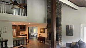 A spacious living area with a high ceiling, a fireplace, and a kitchen in the background. - New 4k sf Mdrn Farmhouse-hot tub-fire pit-games on 3 acres 7 minutes from dwntn. (Asheville)