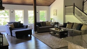 A spacious living room with a high ceiling, a wooden staircase, and large windows. - New 4k sf Mdrn Farmhouse-hot tub-fire pit-games on 3 acres 7 minutes from dwntn. (Asheville)
