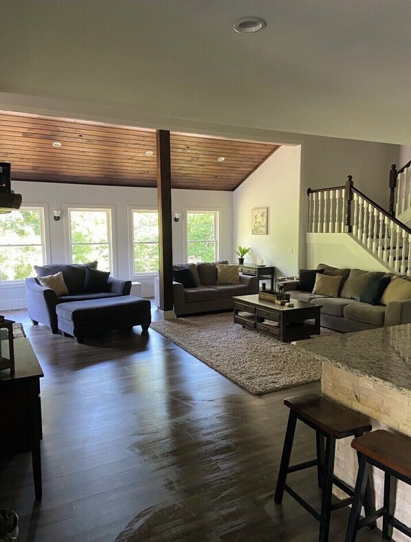 A spacious living room with a high ceiling, a wooden staircase, and large windows.