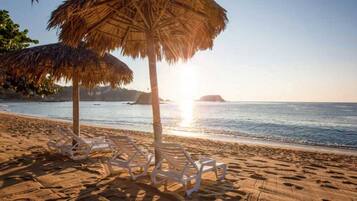 Una playa con sombrillas de paja y sillones blancos.