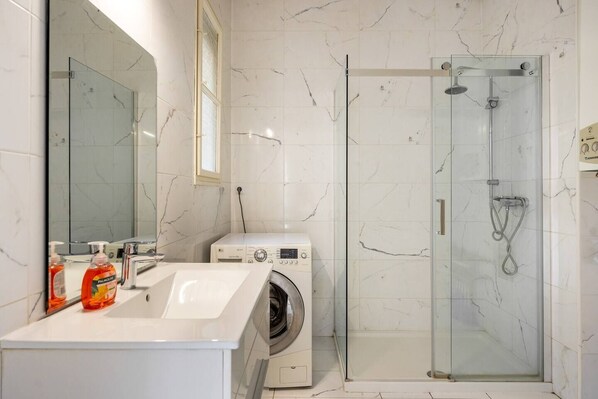 Une salle de bain moderne avec un mur recouvert de carreaux de marbre, une douche en verre, un lave-linge et un lavabo avec un miroir.