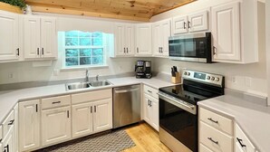 A modern kitchen with white cabinets, a wooden ceiling, and a window providing natural light. - Fun Riverside Cabin: Indoor Games+Hot Tub+Firepit (Ellijay)