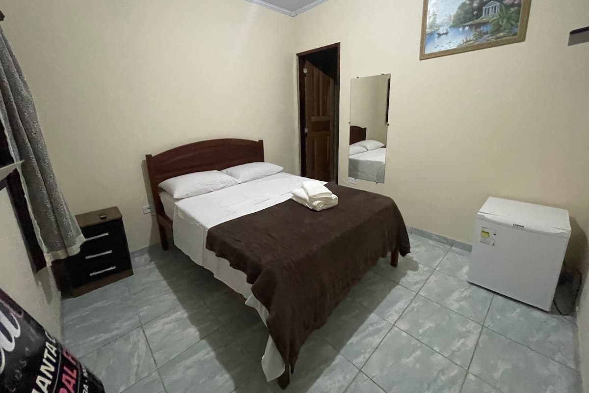 Ein Schlafzimmer mit Bett, Nachttisch, Spiegel, kleinem Kühlschrank und einem Bild an der Wand.