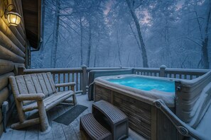 A wooden deck with a hot tub and a chair, surrounded by snow-covered trees.