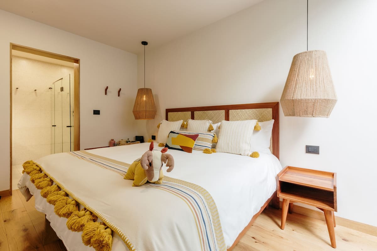 Una cama bien hecha con una colcha a rayas y un peluche encima.  Junto a la cama, una mesita de noche de madera con una lámpara de fibras naturales.