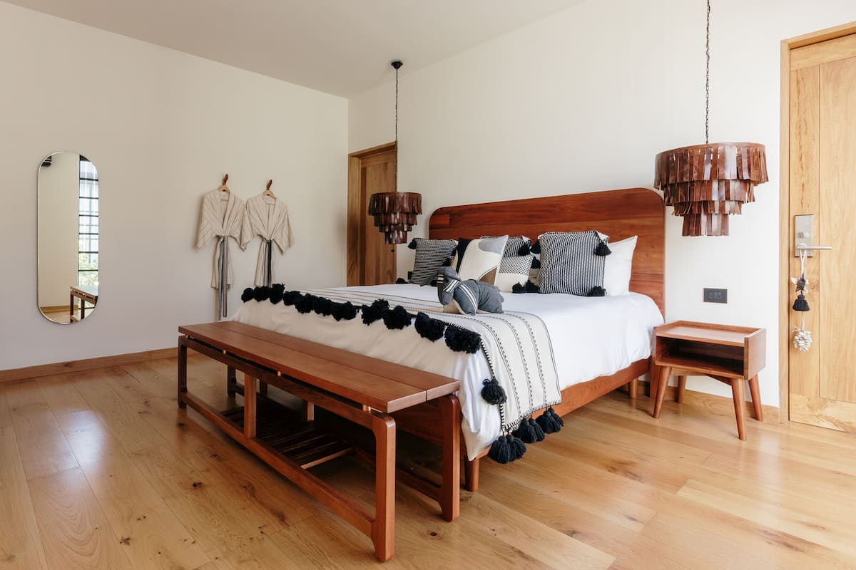 Un dormitorio con una cama de madera, un banco, una mesita de noche, una lámpara colgante y un espejo en la pared.