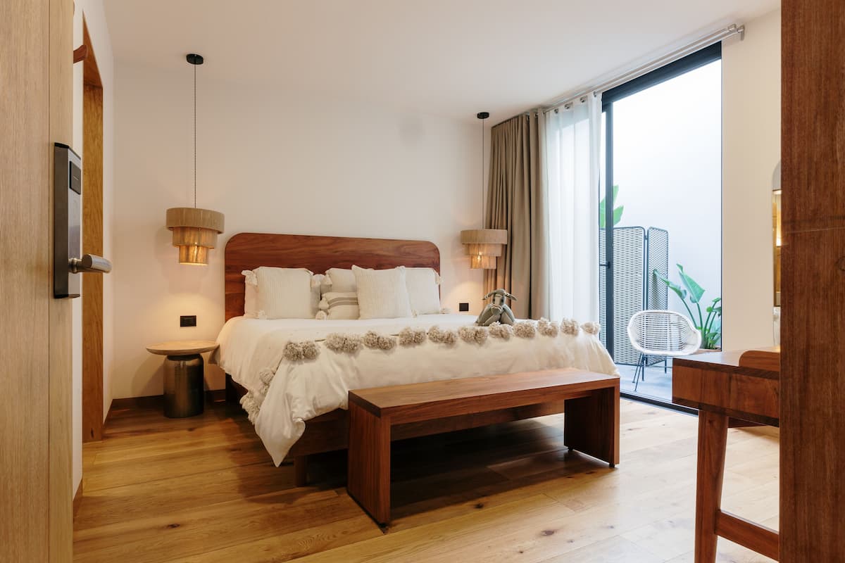 Un dormitorio con cama de madera, mesitas de noche, un banco y vistas a un balcón con plantas.