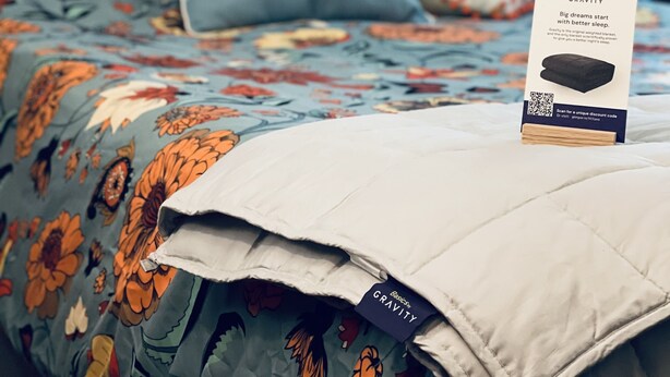 Een net opgemaakt bed met een beddegoed met bloemenprint, een kussen met de tekst 'love you more' en een kussen met het thema zwaartekracht. Een klein bordje met een QR-code staat op het bed.
