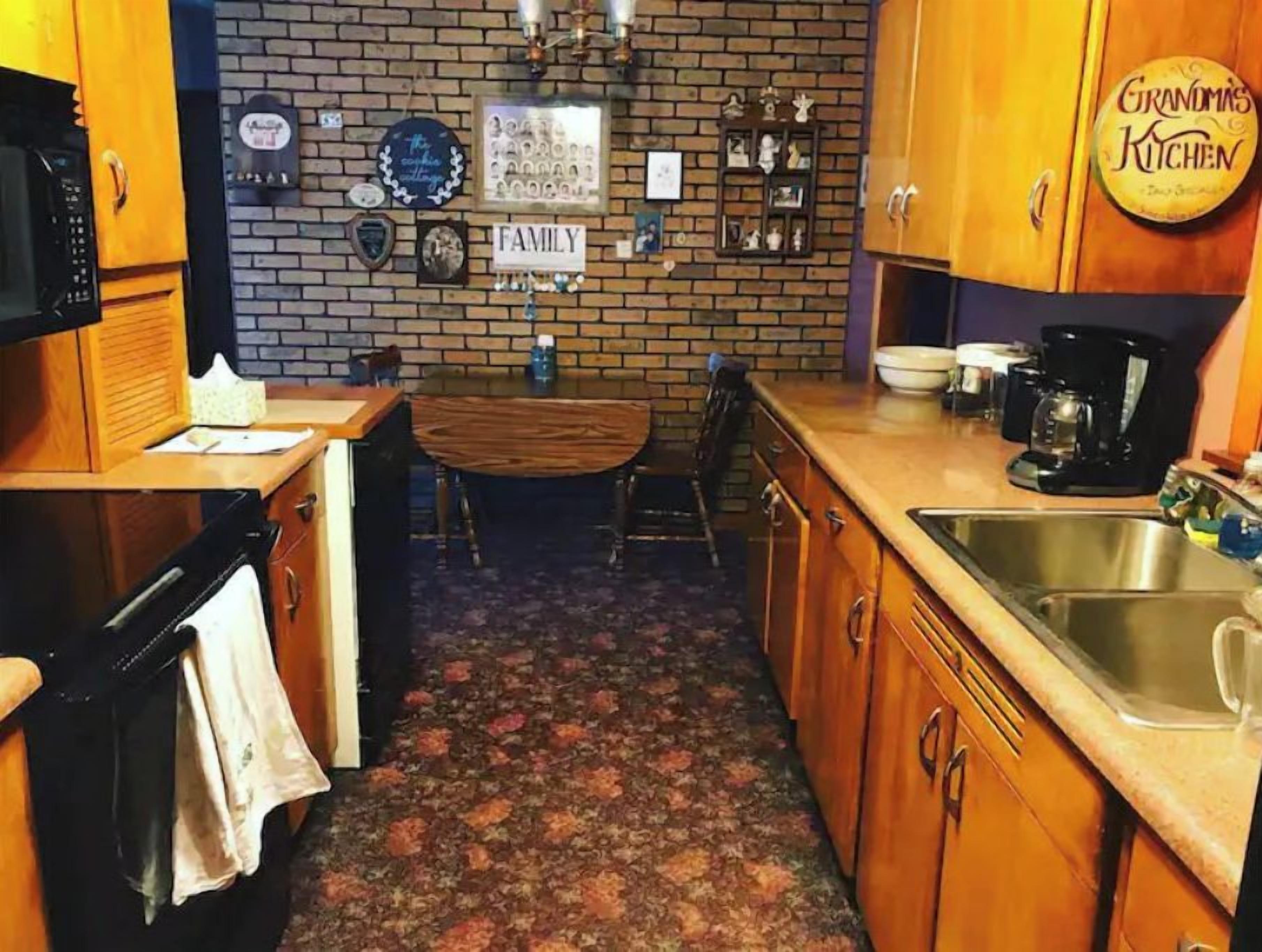 Cuina amb armaris de fusta, paret de maó, cafetera i un rètol que diu "Grandma's Kitchen".