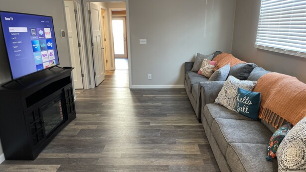 A living room with a flat-screen TV on a black stand, a grey sofa with colorful throw pillows, and a wooden floor.