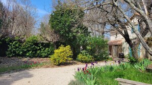 Camí de grava que condueix a un edifici rústic amb teulada de teules, envoltat de vegetació exuberant i plantes amb flors.