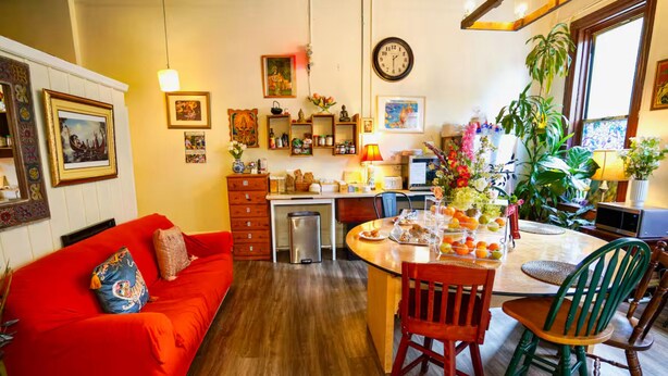 A cozy living room with a red sofa, a dining area with a wooden table and chairs, and a kitchen with wooden cabinets and a sink.