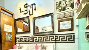 A bathroom with two sinks, mirrors, and a tiled backsplash. There are shelves with toiletries and a trash can.