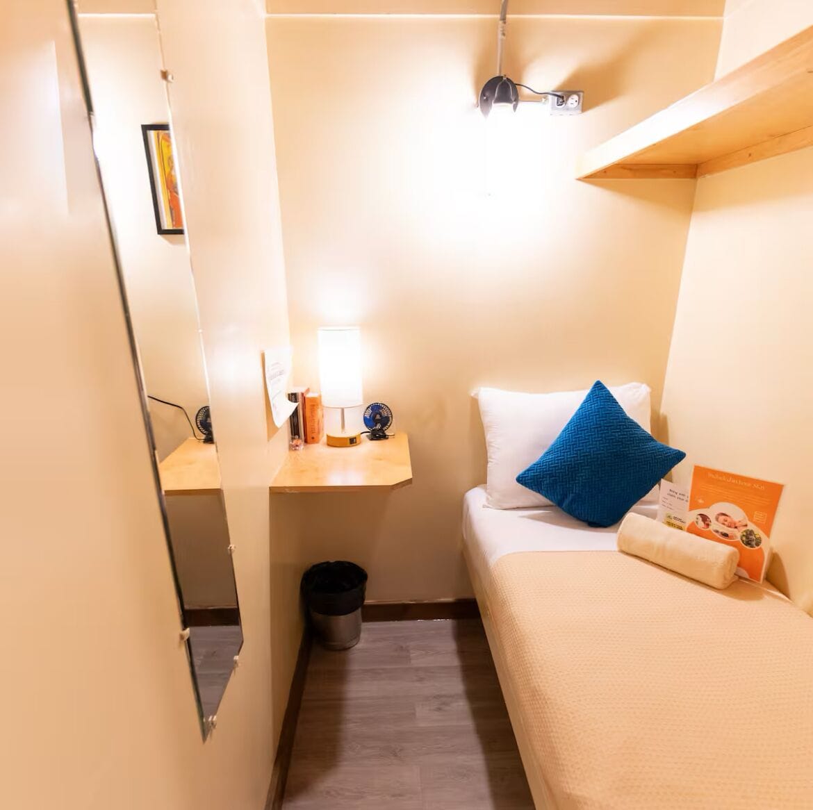Une petite pièce, de taille modeste, comprenant un lit, une table de chevet avec une lampe, un ventilateur et une poubelle.