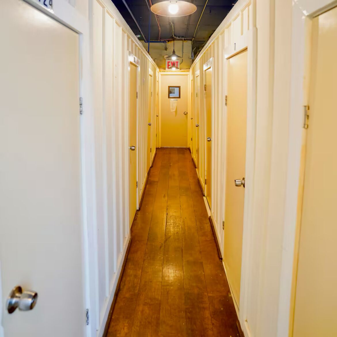A narrow hallway with doors on both sides, a wooden floor, and an exit sign above.