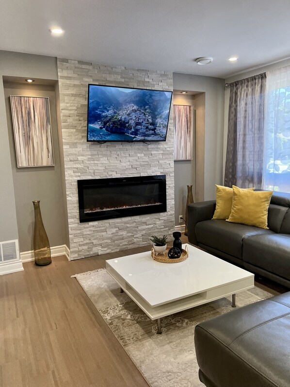 A modern living room with a stone fireplace, a flat-screen TV mounted above it, a black leather sofa with yellow cushions, and a white coffee table.