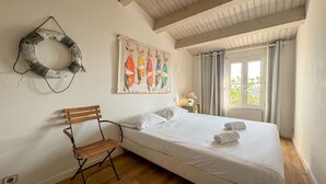 Un dormitorio con una cama, una silla de madera, un cuadro colgado en la pared, una ventana con cortinas y un adorno de pared.